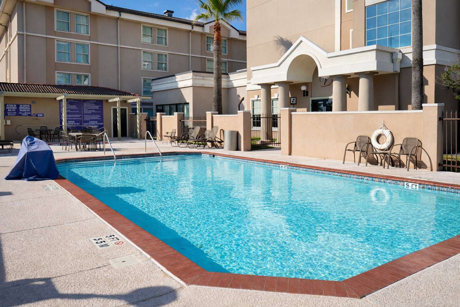 Outdoor pool with lounge chairs and shaded patio tables beside the hotel's arched entrance and palm trees