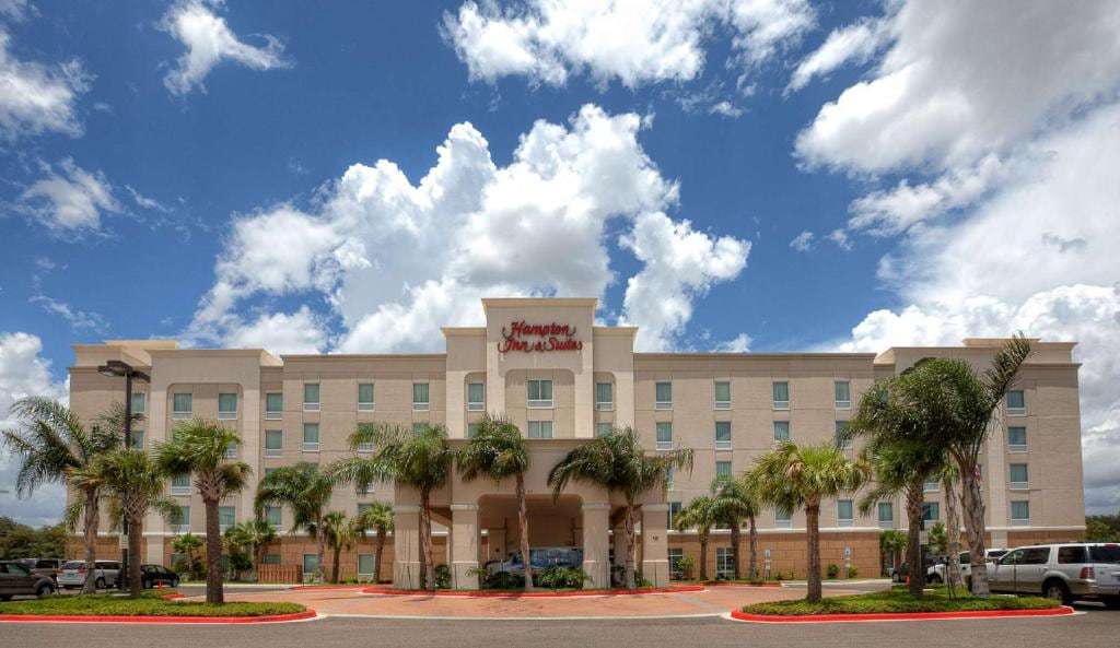Hampton Inn & Suites front facade with palm-lined entrance drive under a blue sky with clouds