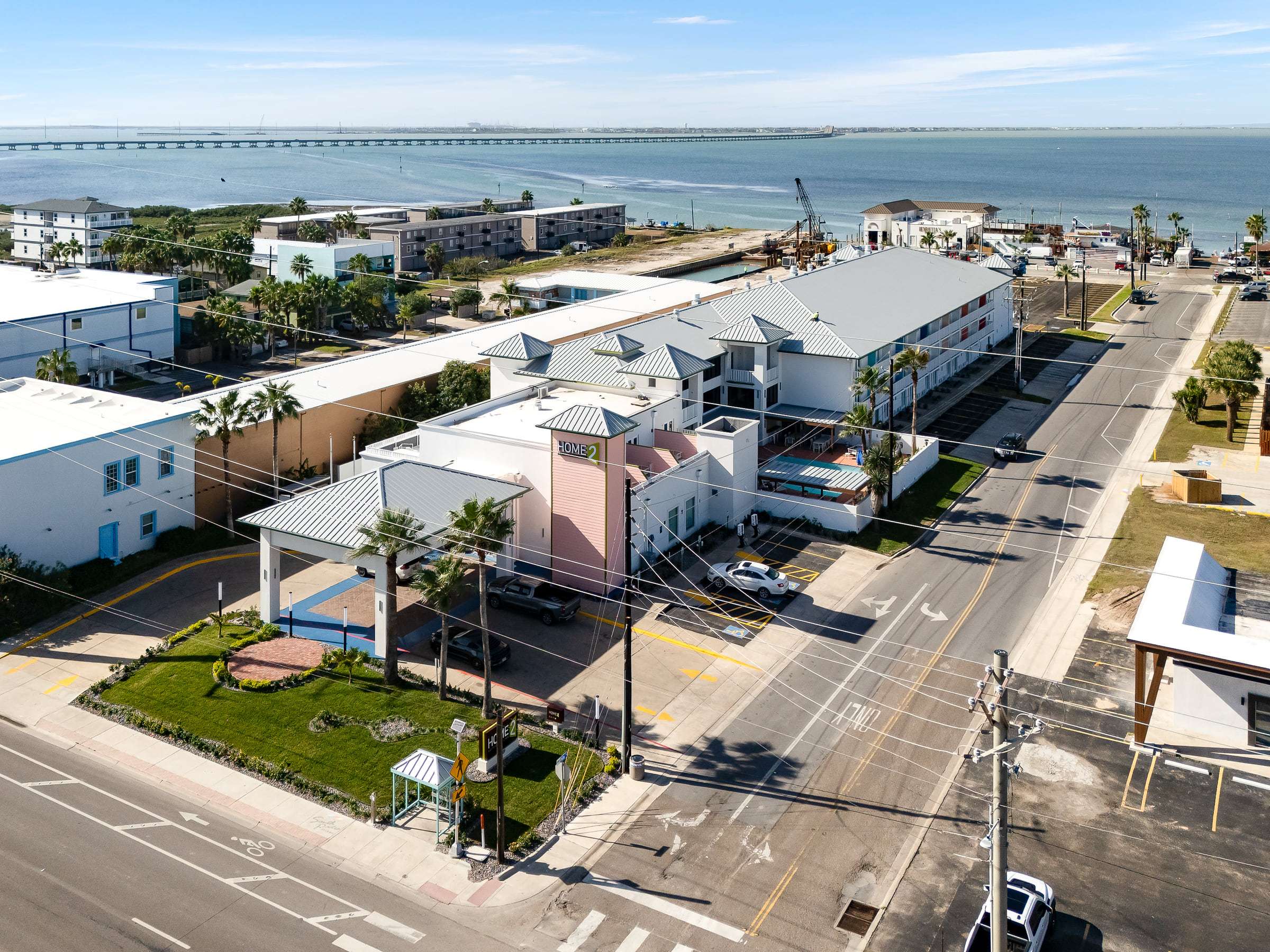 Aerial view of the hotel on Padre Blvd with its pink Home2 tower, the bay and the causeway behind