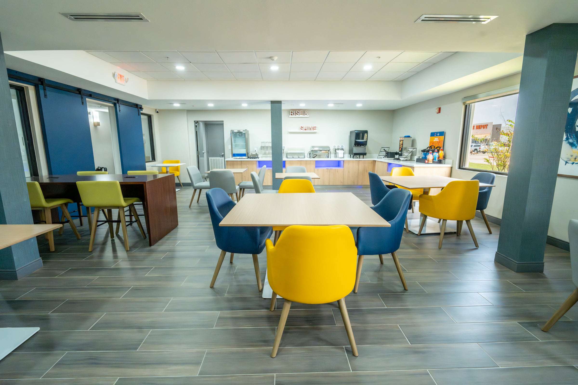Breakfast room with yellow and blue chairs at wood tables and a buffet counter under a Rise and Shine sign