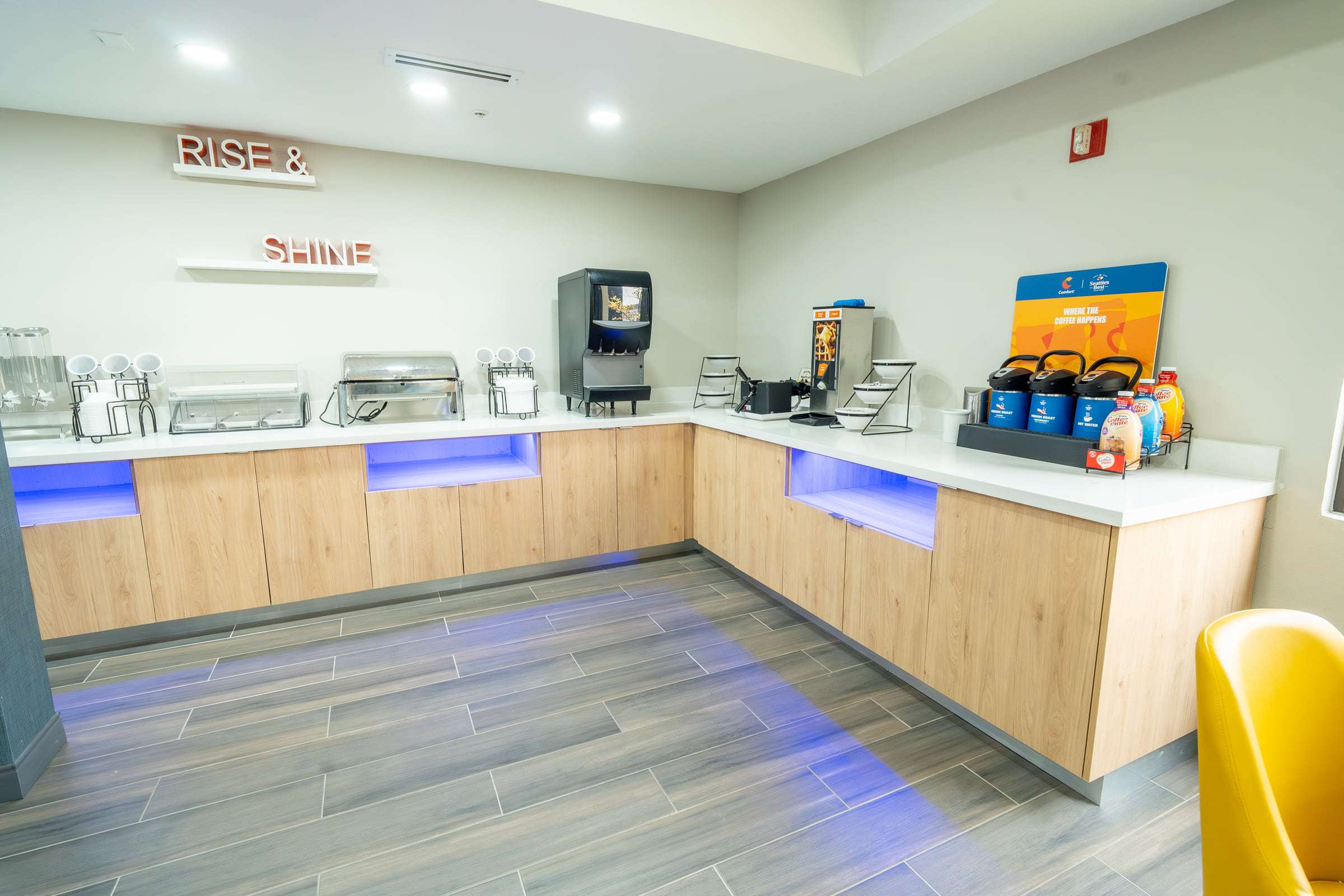 Breakfast buffet counter with chafing dishes, beverage machines and coffee airpots below a Rise and Shine sign