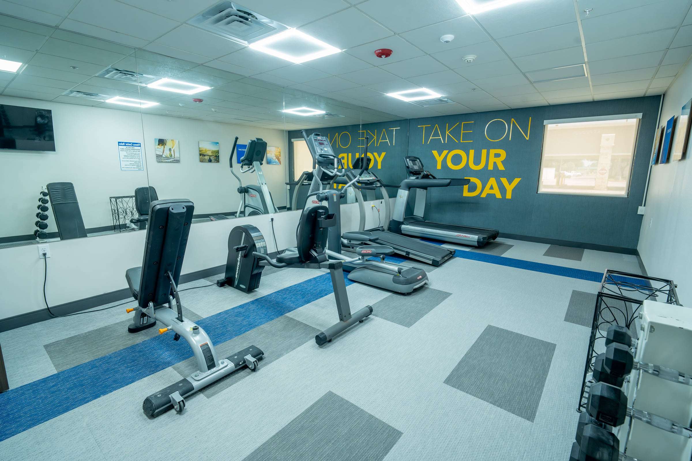 Fitness room with treadmills, ellipticals, a recumbent bike, dumbbell rack and a wall reading Take On Your Day
