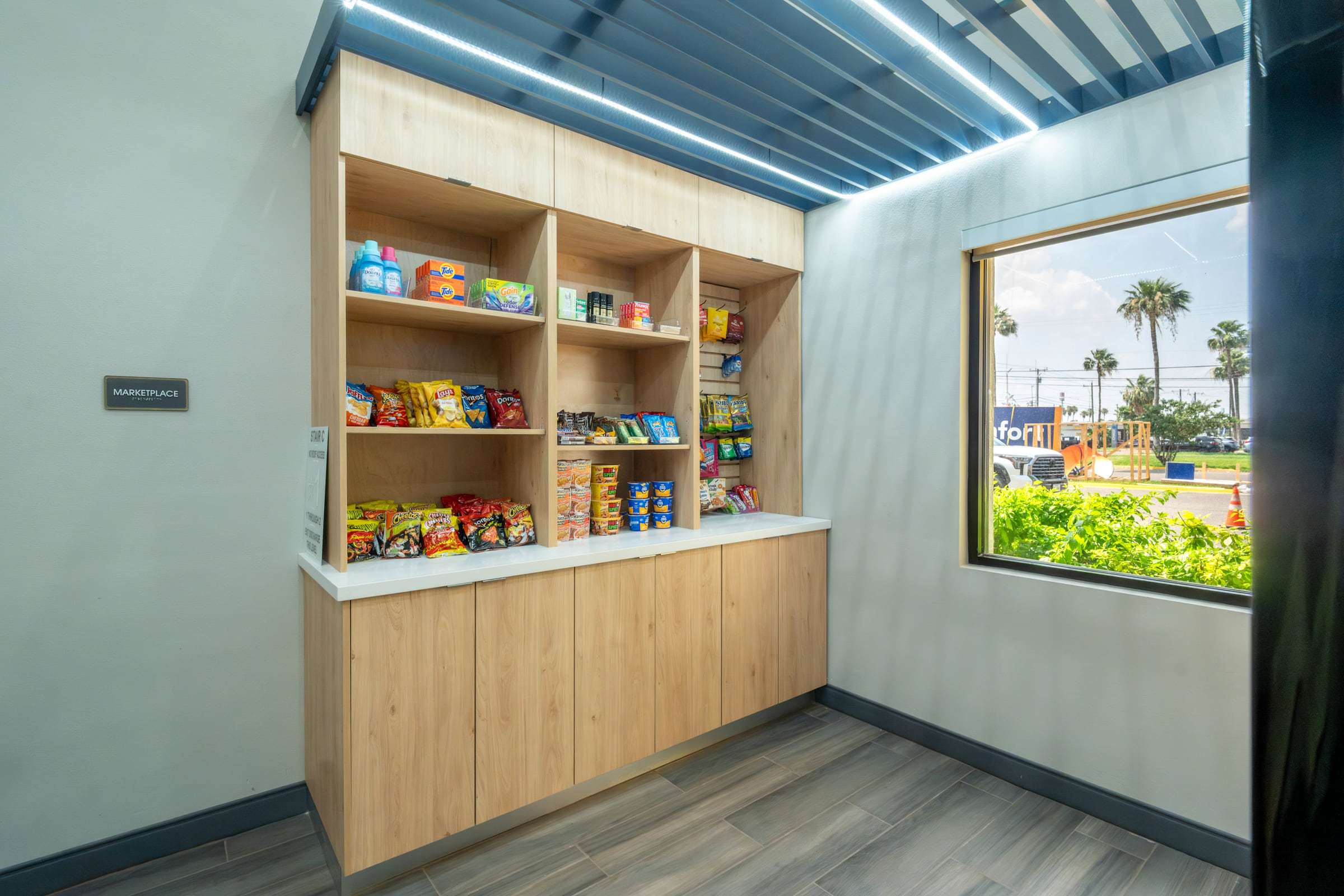 Wood shelving stocked with snacks and toiletries for sale beside a window with palm trees outside