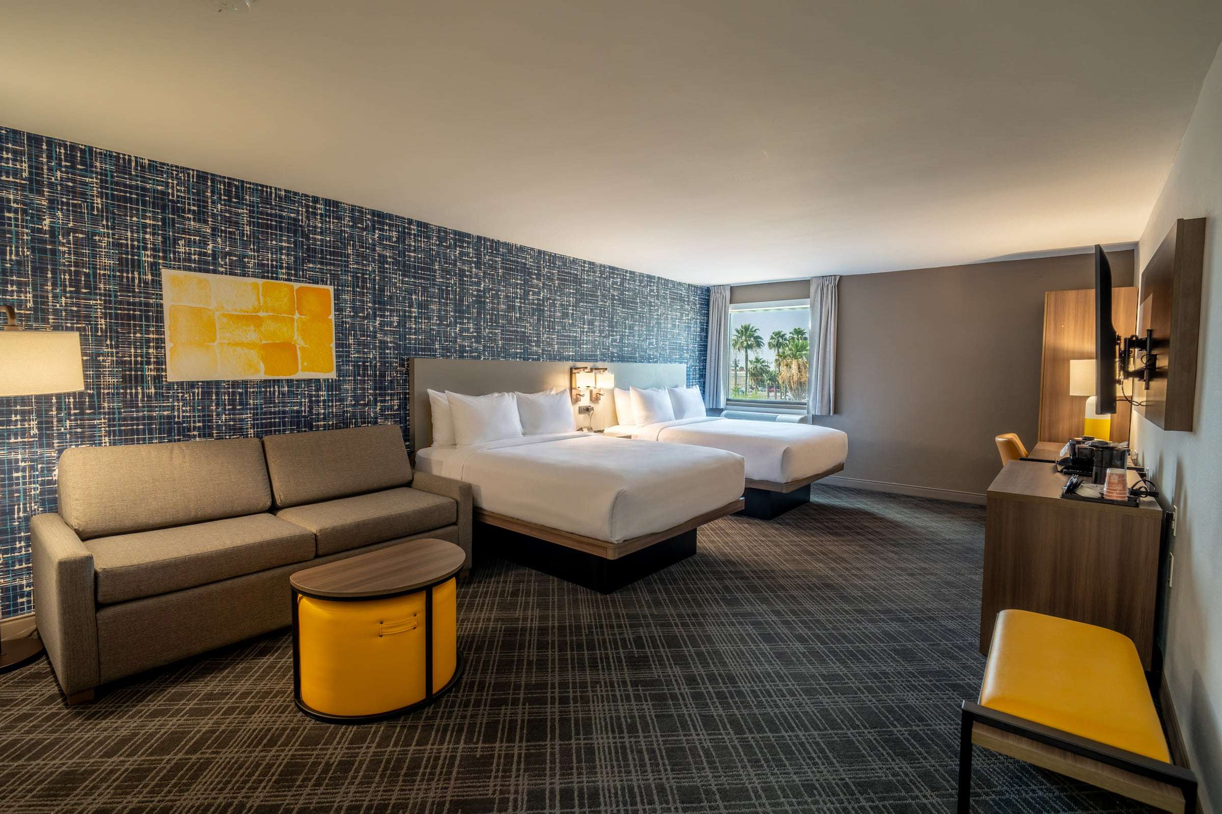 Guest room with two queen beds, a gray sofa, a round yellow side table and a wall-mounted TV