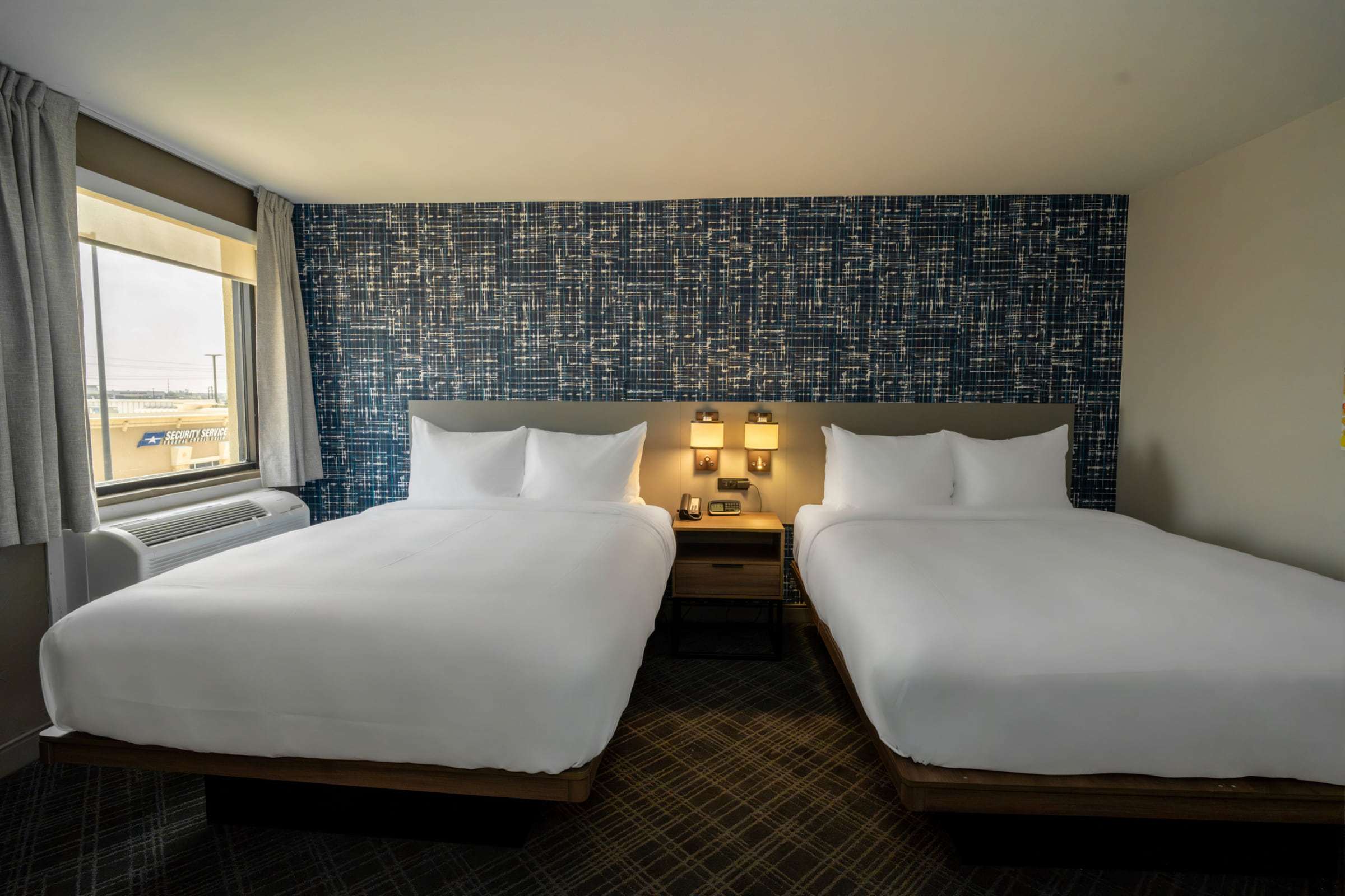 Two queen beds with white bedding set against a blue patterned accent wall, beside a window and air-conditioning unit