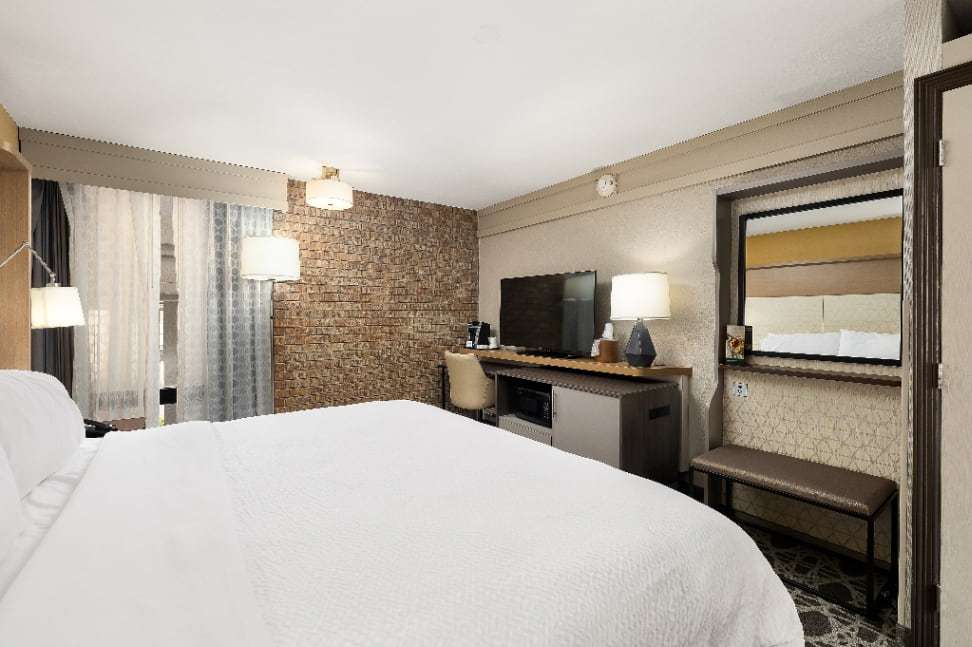 Guest room with large bed, TV console with microwave, wall mirror and upholstered bench