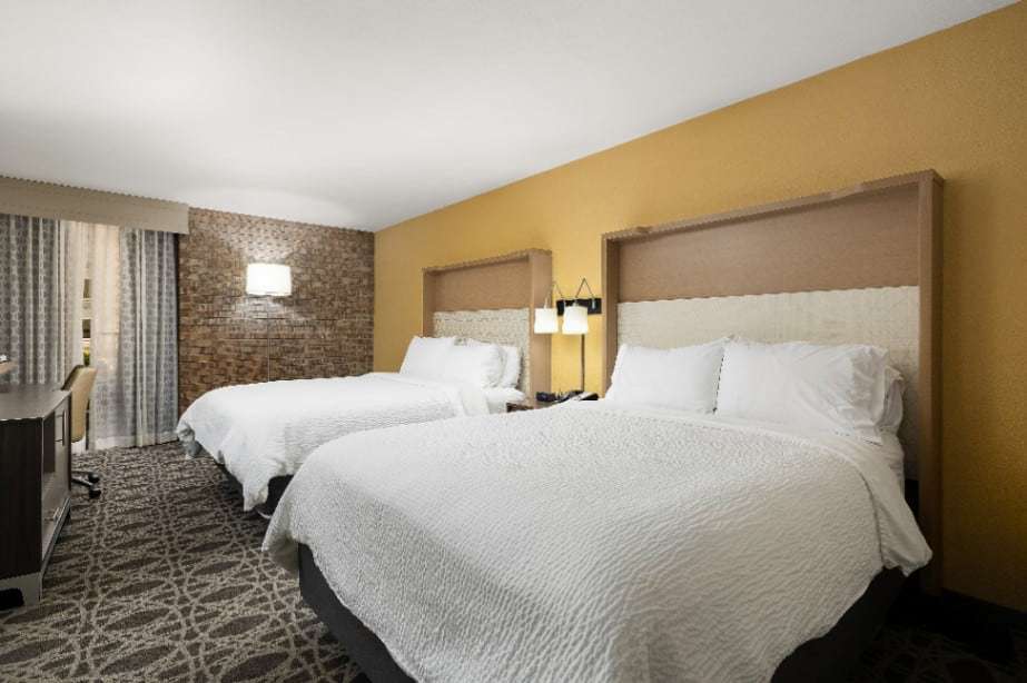 Two queen beds with tall upholstered headboards, brick wall and desk by the window