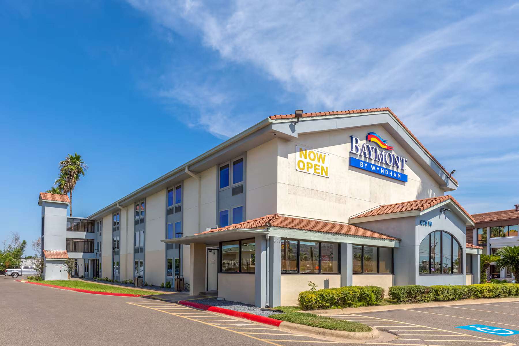 Hotel exterior by day with Baymont by Wyndham sign, tile-roof entrance and palm trees