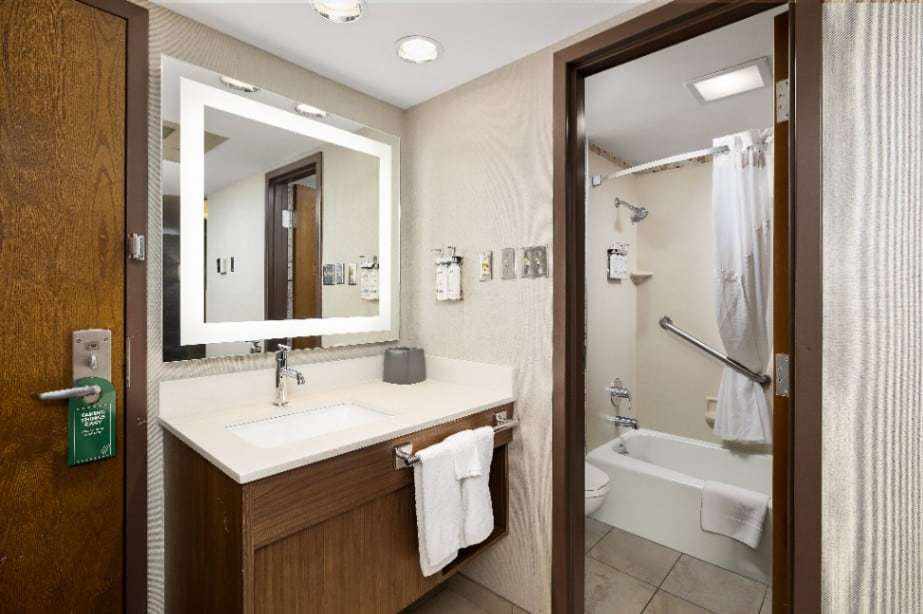 Vanity with lighted mirror next to a bathroom with tub-shower and grab bar