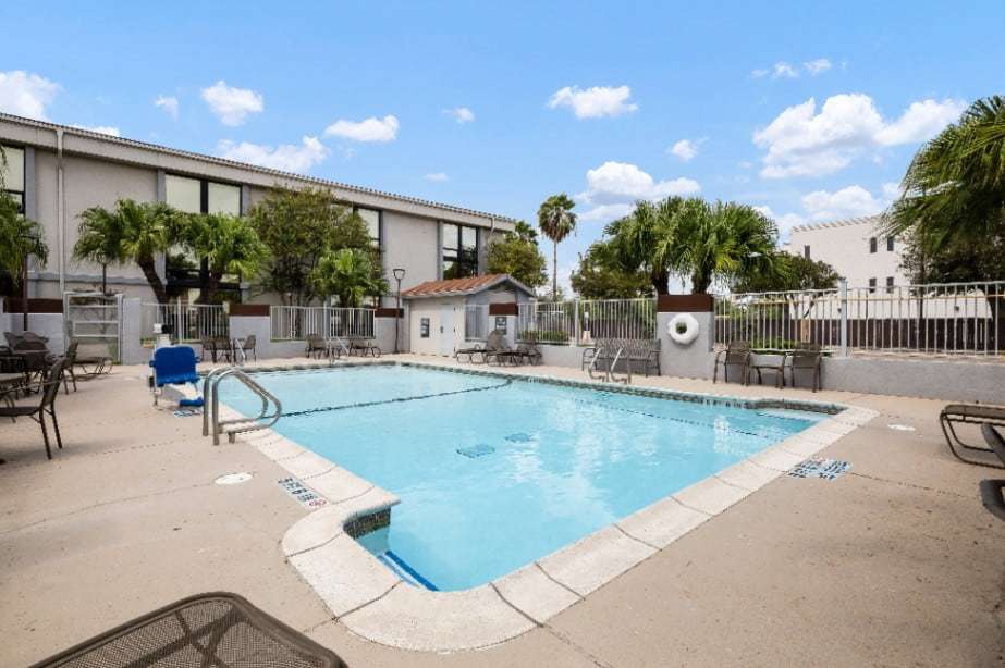 Fenced outdoor pool with lounge chairs and palm trees beside a two-story hotel wing