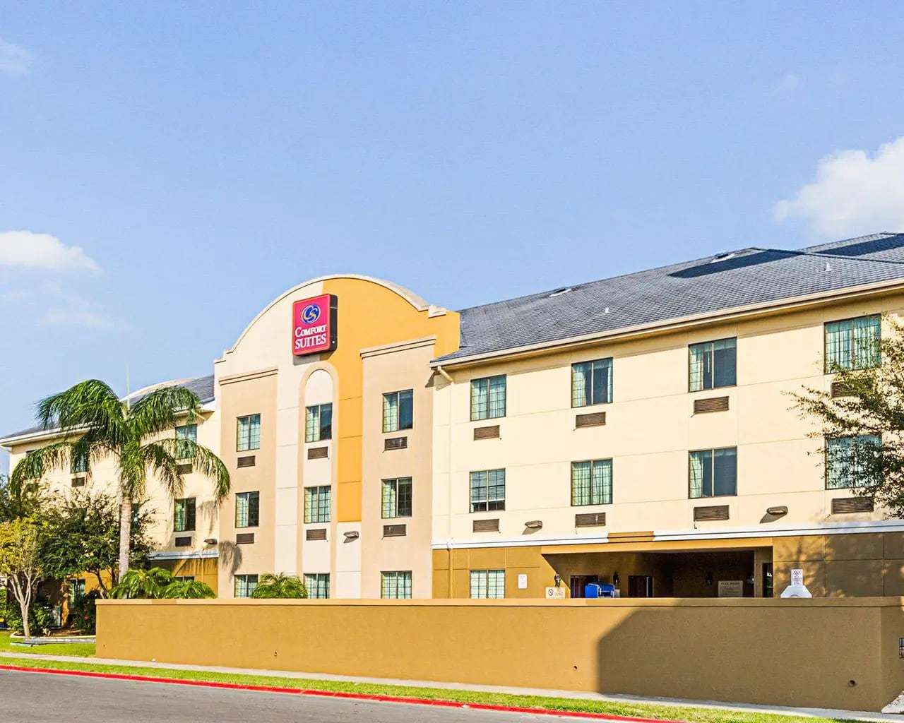 Three-story hotel facade with Comfort Suites sign on an arched tower, palm trees and street wall